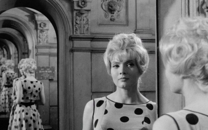 A black and white film still of a woman (Anna Karina) with blond hair and a polka dot, sleeveless dress, looking at reflections of herself and an archway in a series of mirrors.