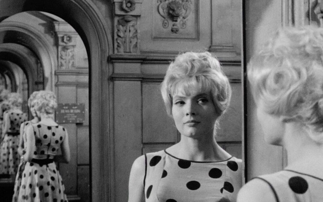 A black and white film still of a woman (Anna Karina) with blond hair and a polka dot, sleeveless dress, looking at reflections of herself and an archway in a series of mirrors.