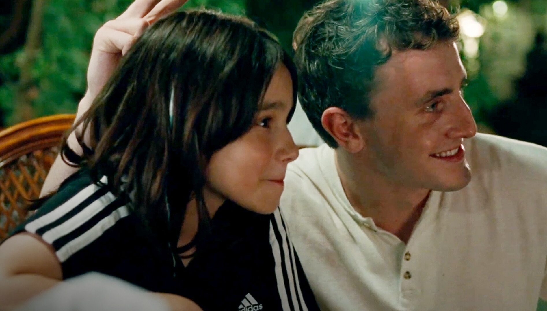 A girl with long brown hair sitting next to a man with blond hair, who is holding up rabbit ears behind her head