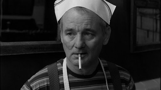 Black and white film still of a middle-aged white man (Bill Murray) with a cook's hat, an apron, and a cigarette hanging out of his mouth.