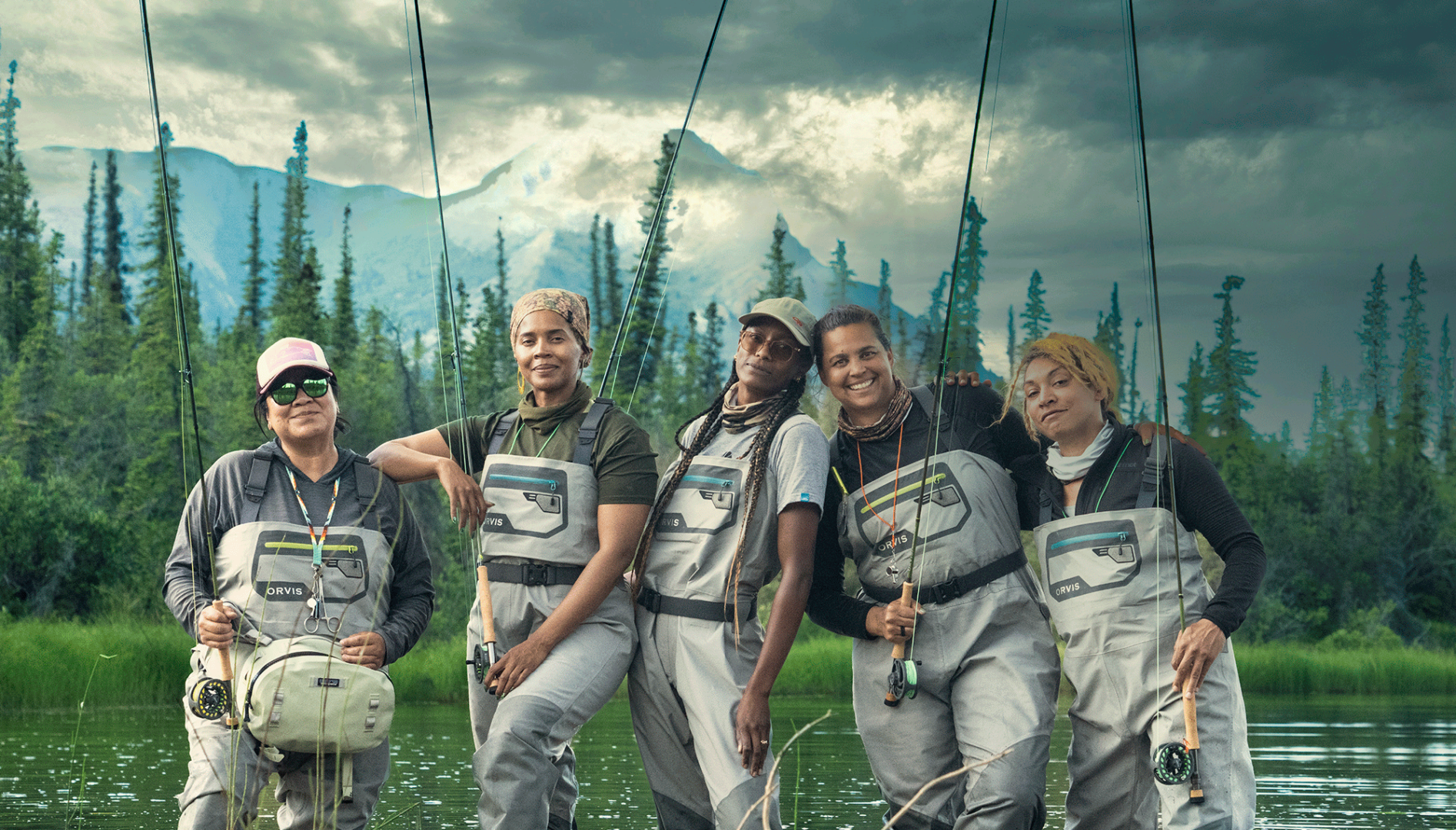 Five BIPOC women standing in water, wearing waders and holding fishing rods, with trees and mountains behind them.