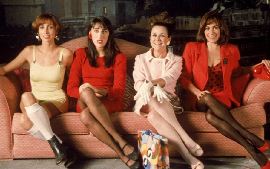 Four women sitting on a pink couch. All of their legs are crossed in the same direction.