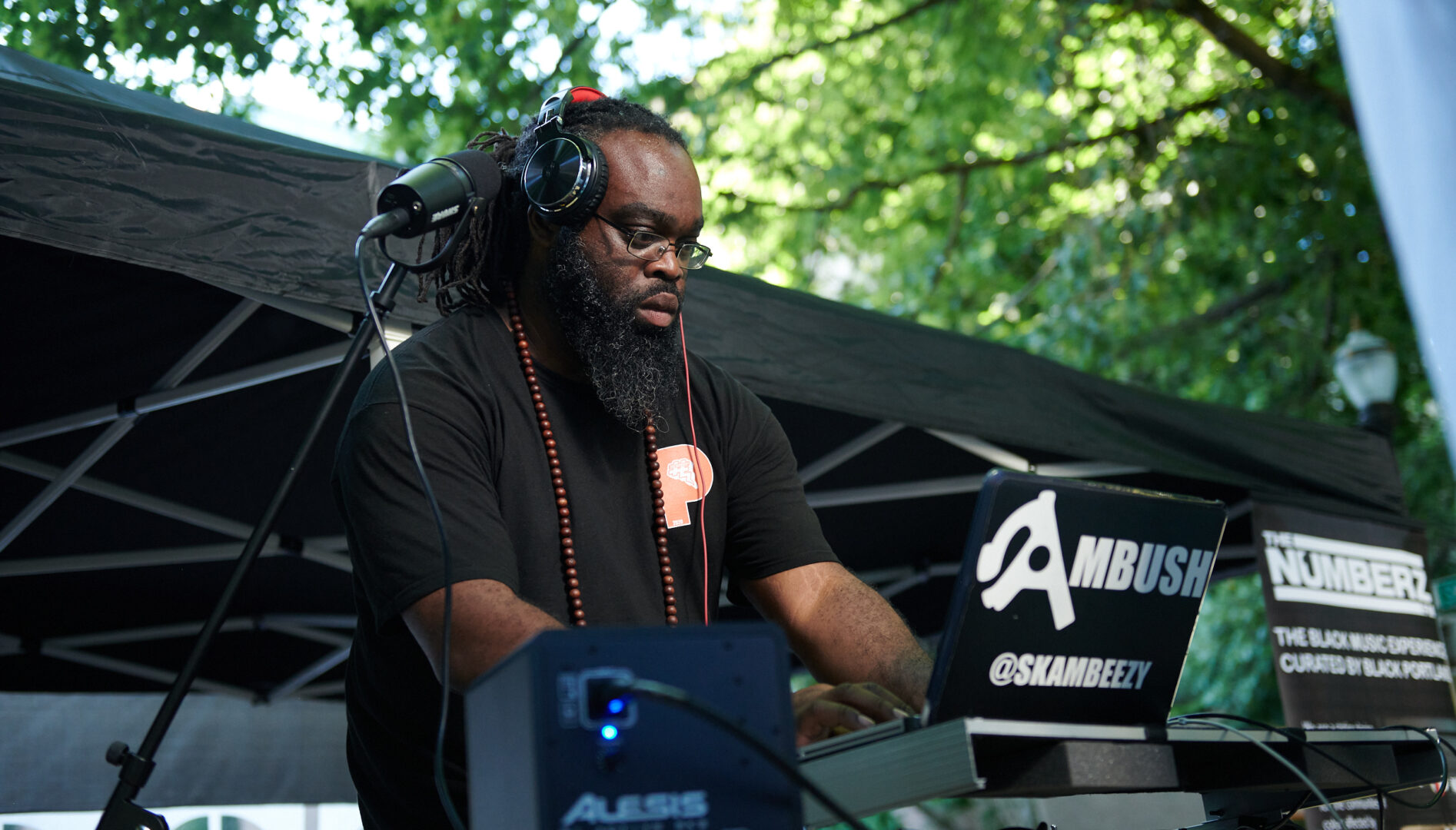 A Black man with dreadlocks, wearing eyeglasses, with headphones on and turntables in front of him