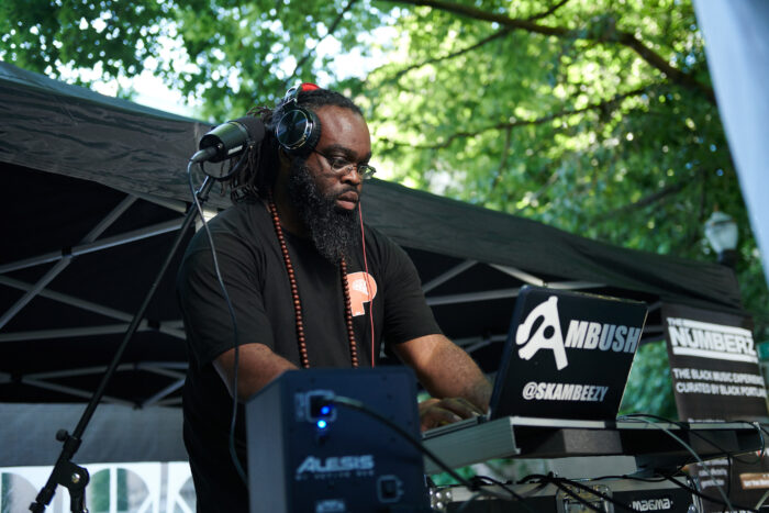 A Black man with dreadlocks, wearing eyeglasses, with headphones on and turntables in front of him
