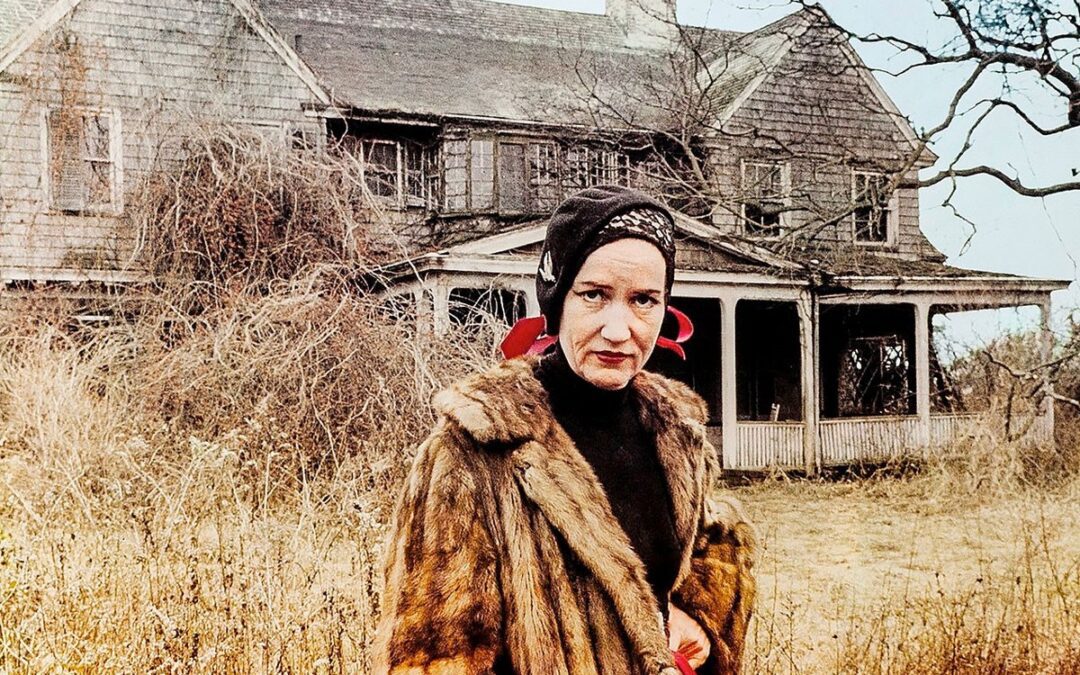 A woman in a fur coat and a black headscarf standing in front of a large rundown house with dead plants and grass behind her