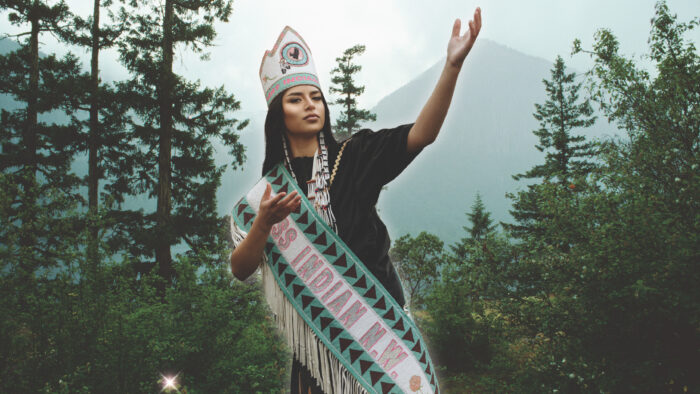 A young Native woman wearing a sash and tall hat saying "Miss Indian N.W." in front of trees and a mountain.