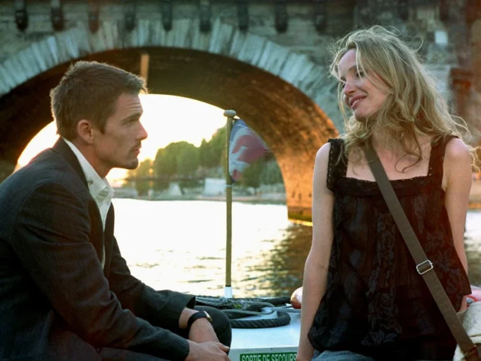 Film still of a man (Ethan Hawke) and a woman with long blond hair (Julie Delpy) sitting in front of a bridge and a river.