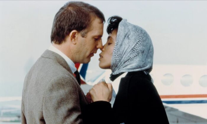A white man with short brown hair (Kevin Costner) about to kiss a Black woman with a light blue headscarf on (Whitney Houston)