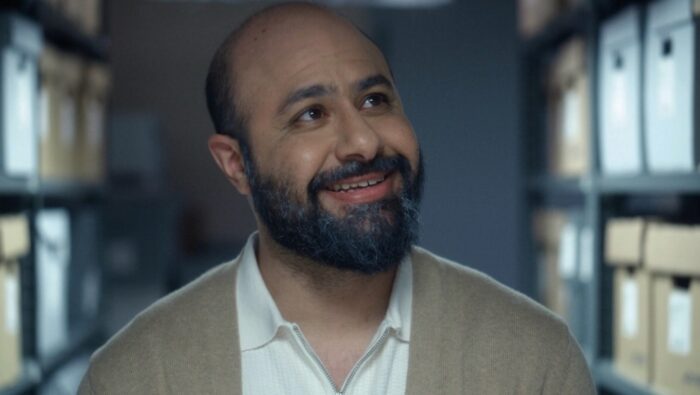 A man with a beard and mustache looking up and smiling to the right of the viewer. Rows of boxes on shelves are behind him.