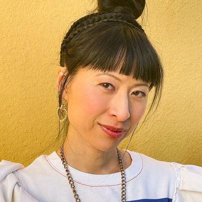 Photo of a woman with her dark hair up in a bun, standing against a yellow wall.