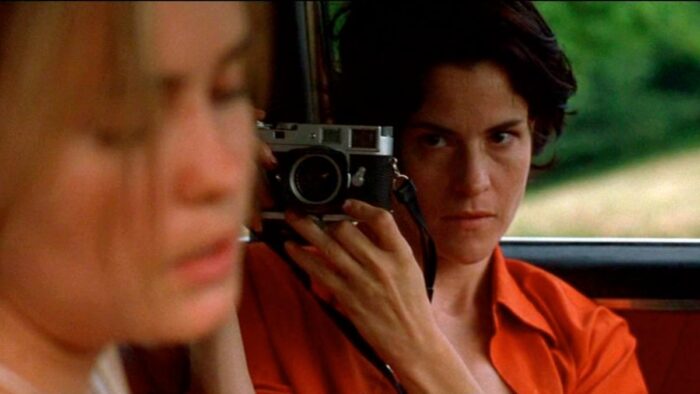 Two women in the front seat of a car. One woman is holding up a camera towards the other, who has her eyes closed