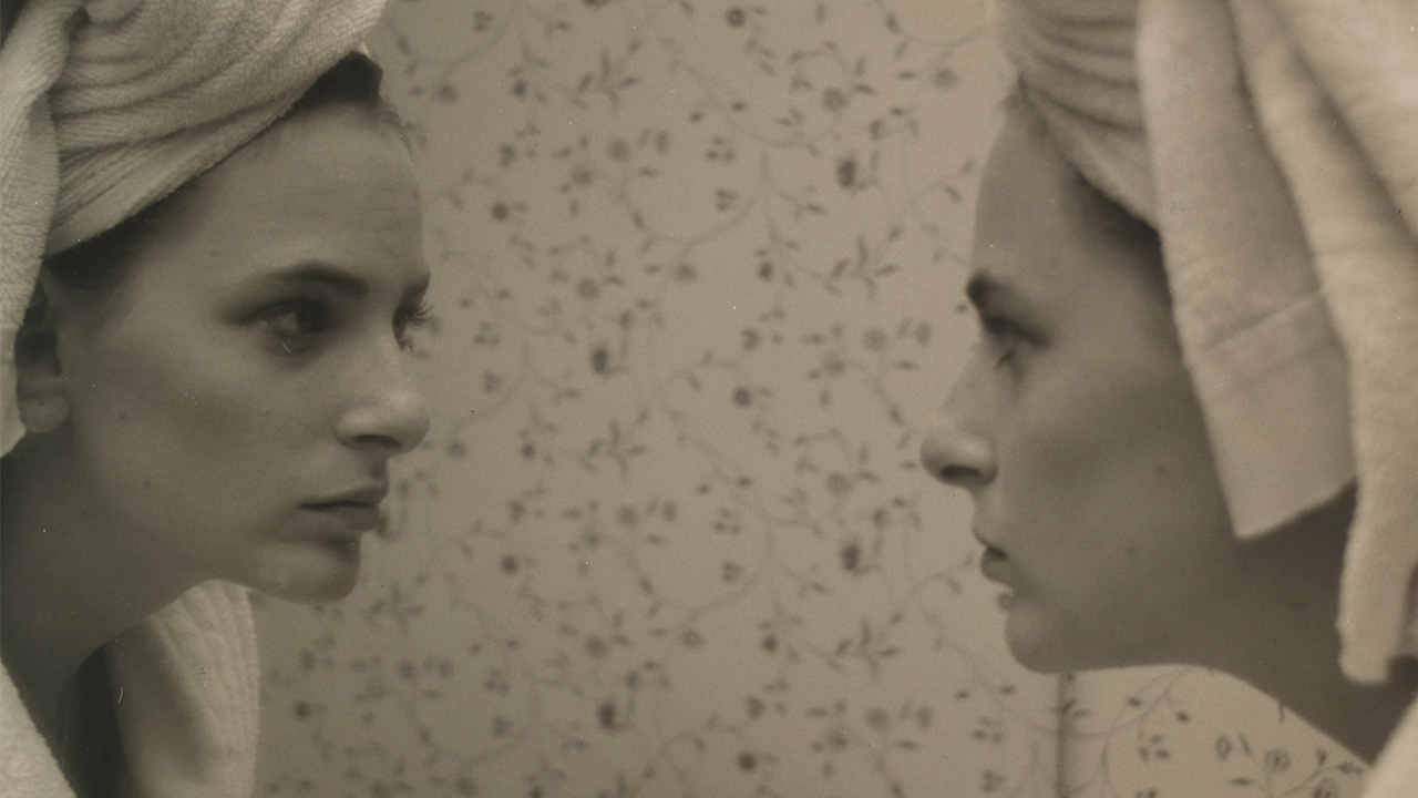 A sepia-toned film still of a woman with a towel wrapped around her head looking at her reflection in a mirror