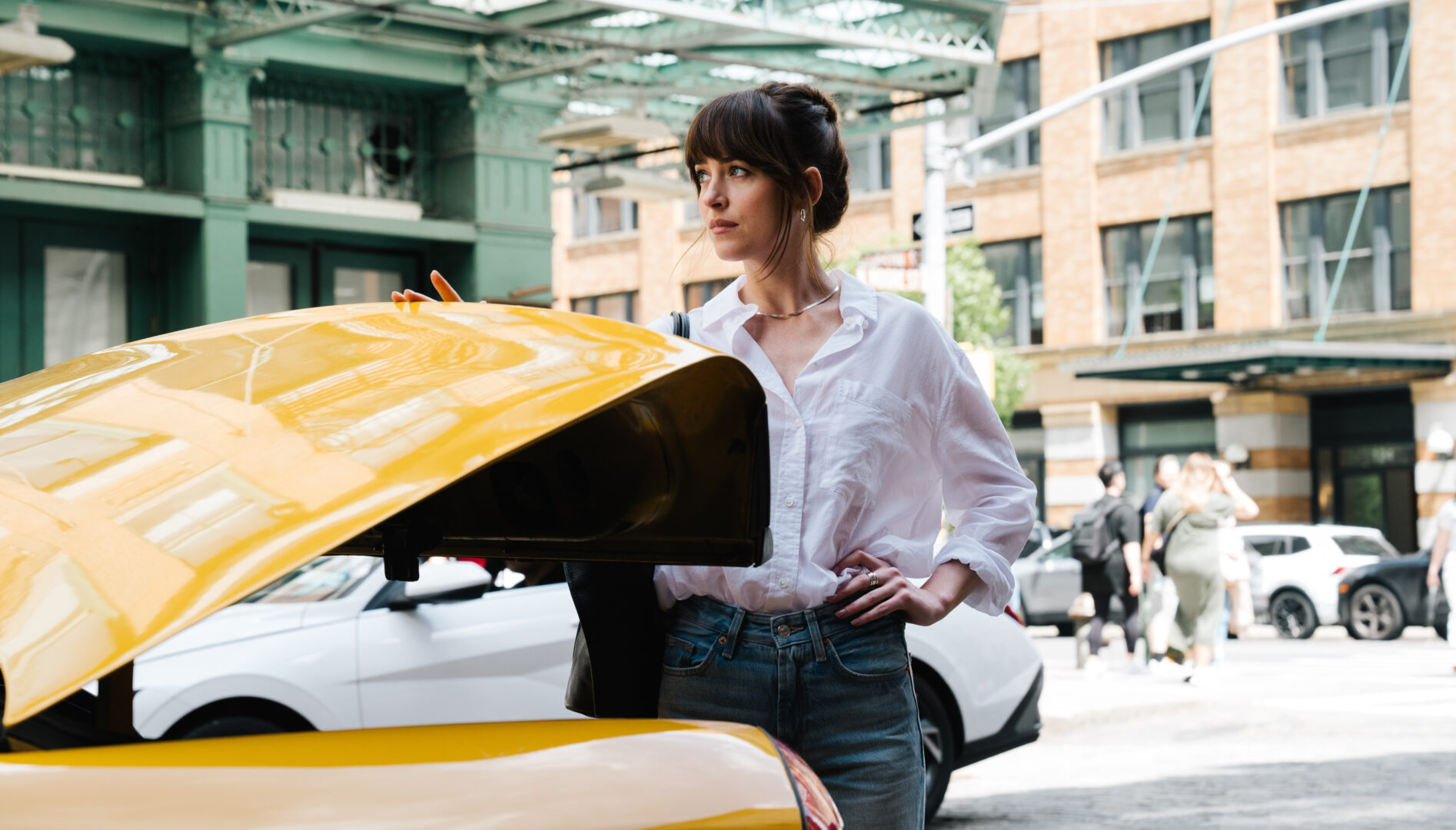 A woman with dark brown hair standing in a city street next to the partially opened trunk of a yellow taxi