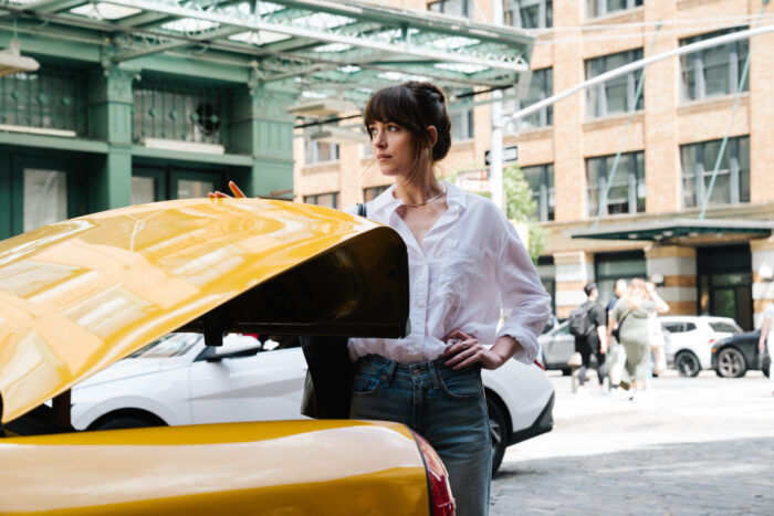 A woman with dark brown hair standing in a city street next to the partially opened trunk of a yellow taxi