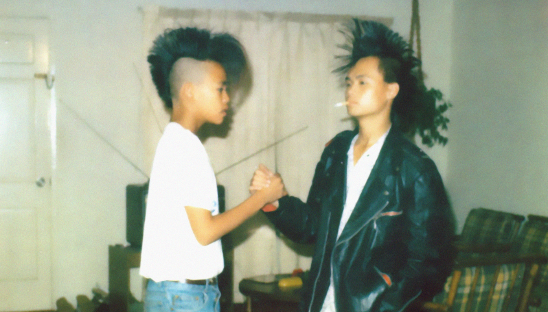 A young boy in a white t-shirt and a young man in a white t-shirt and black leather jacket with a cigarette hanging out of his mouth. They both have mohawks and are clasping opposite hands in a handshake.