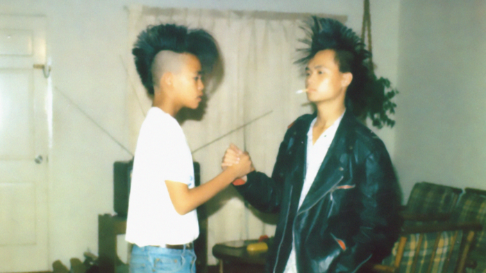 A young boy in a white t-shirt and a young man in a white t-shirt and black leather jacket with a cigarette hanging out of his mouth. They both have mohawks and are clasping opposite hands in a handshake.