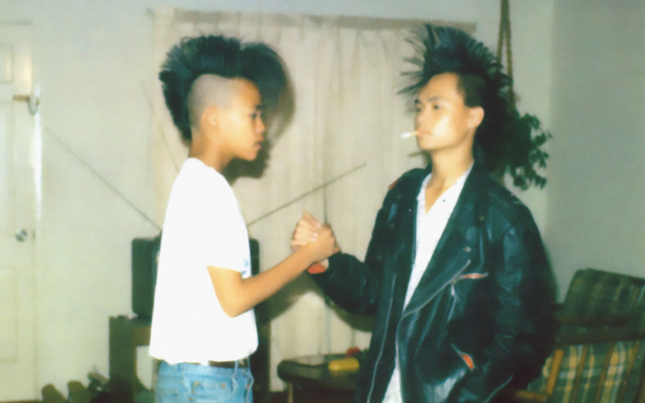 A young boy in a white t-shirt and a young man in a white t-shirt and black leather jacket with a cigarette hanging out of his mouth. They both have mohawks and are clasping opposite hands in a handshake.