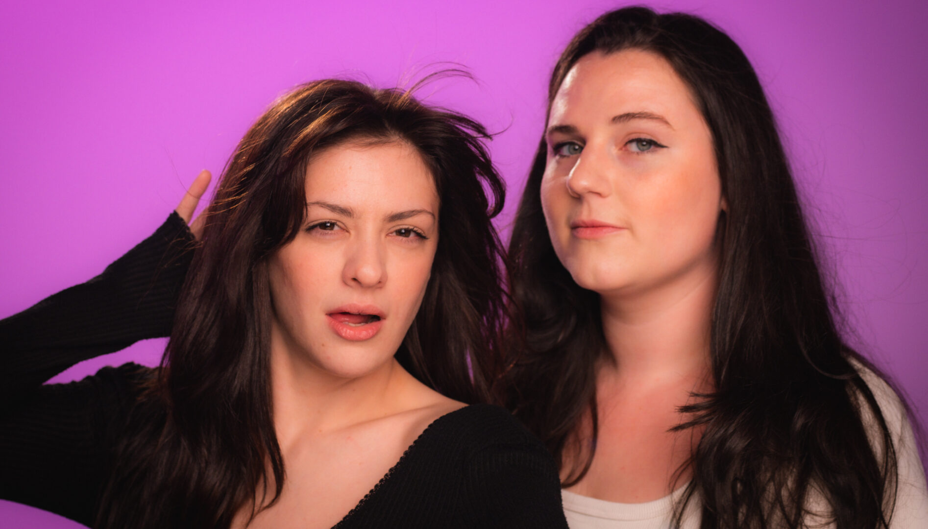 Two young women with long brown hair standing against a purple backdrop