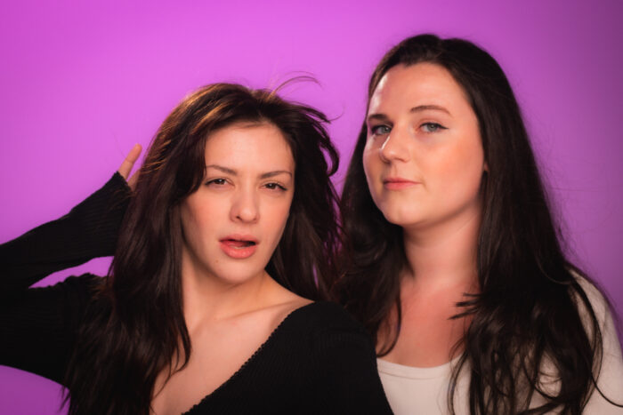 Two young women with long brown hair standing against a purple backdrop