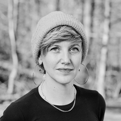 Black and white photo of a woman with short blond hair wearing a beanie with trees behind her.