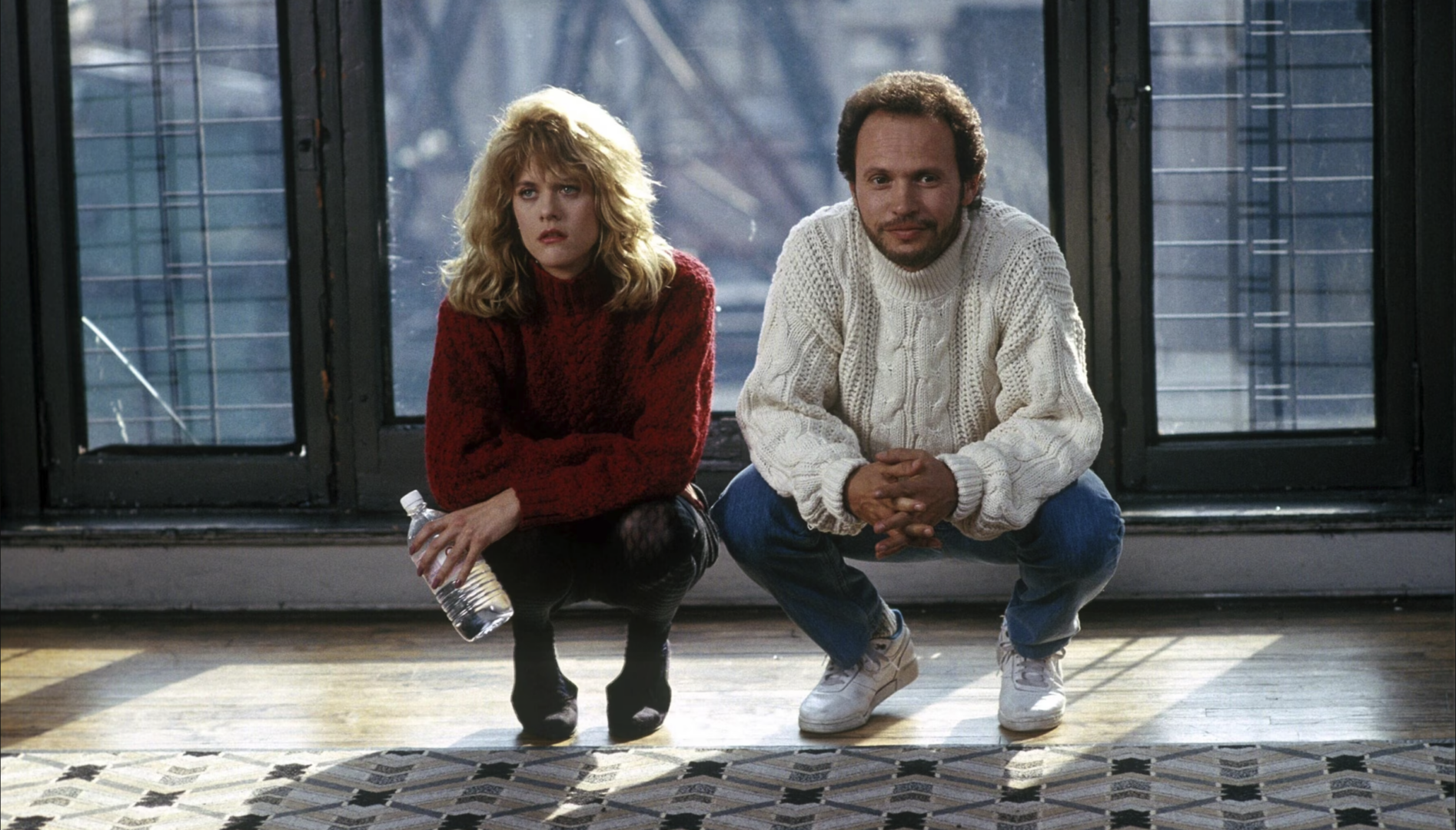 A woman with long curly blonde hair and a man with short dark hair crouched next to each other in front of a patterned rug, with large windows behind them