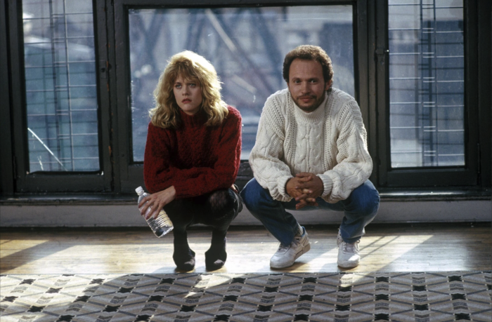 A woman with long curly blonde hair and a man with short dark hair crouched next to each other in front of a patterned rug, with large windows behind them