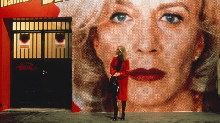 A blond woman in a red coat stands in front of a large image of the face of a woman who has curly blond hair.