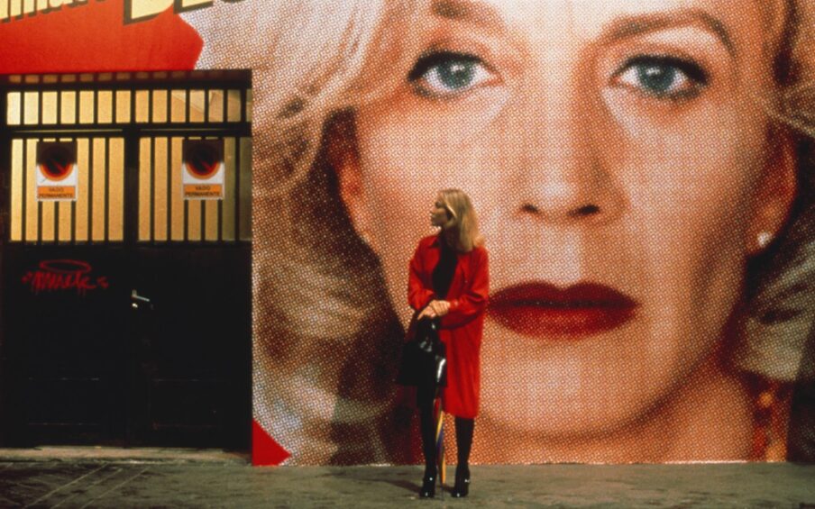 A blond woman in a red coat stands in front of a large image of the face of a woman who has curly blond hair.