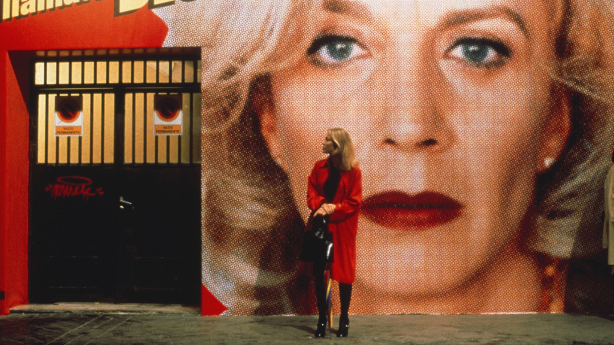 A blond woman in a red coat stands in front of a large image of the face of a woman who has curly blond hair.
