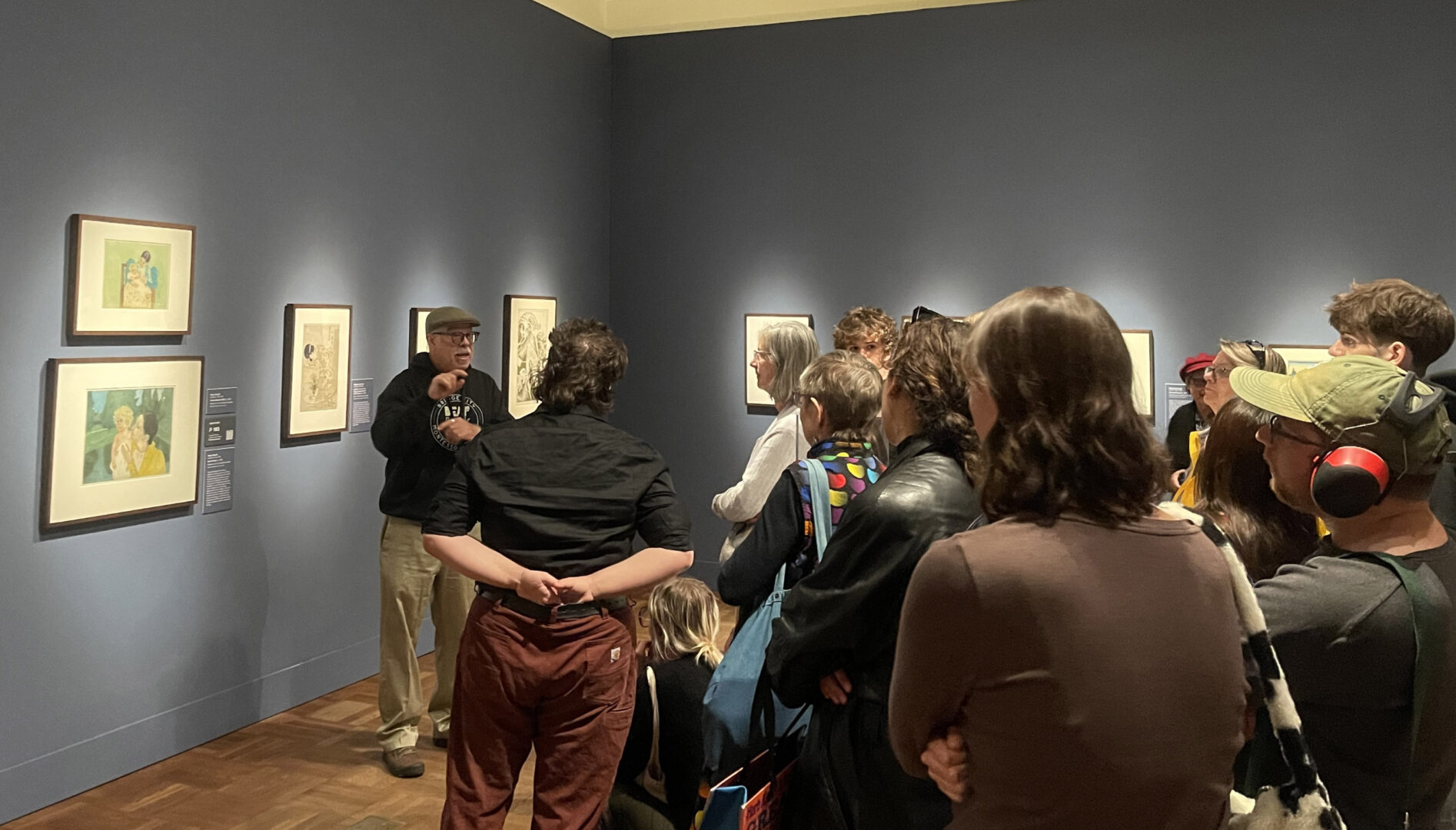 A group of people standing in a gallery with art on the walls, listening to a presenter.