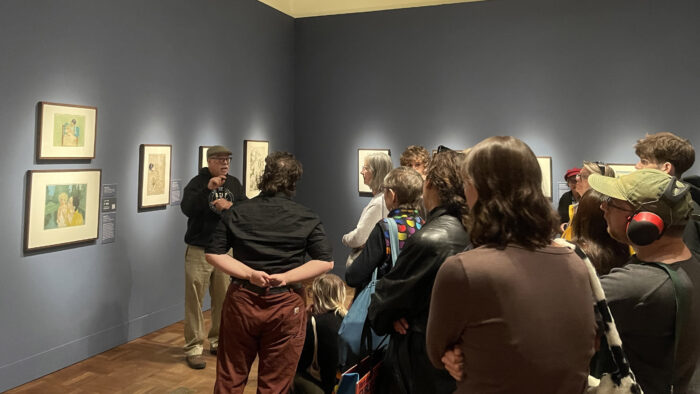 A group of people standing in a gallery with art on the walls, listening to a presenter.