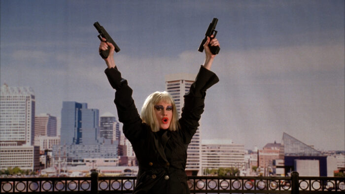 A woman wearing a blond wig and a black trenchcoat holding two guns up in the air against a cityscape