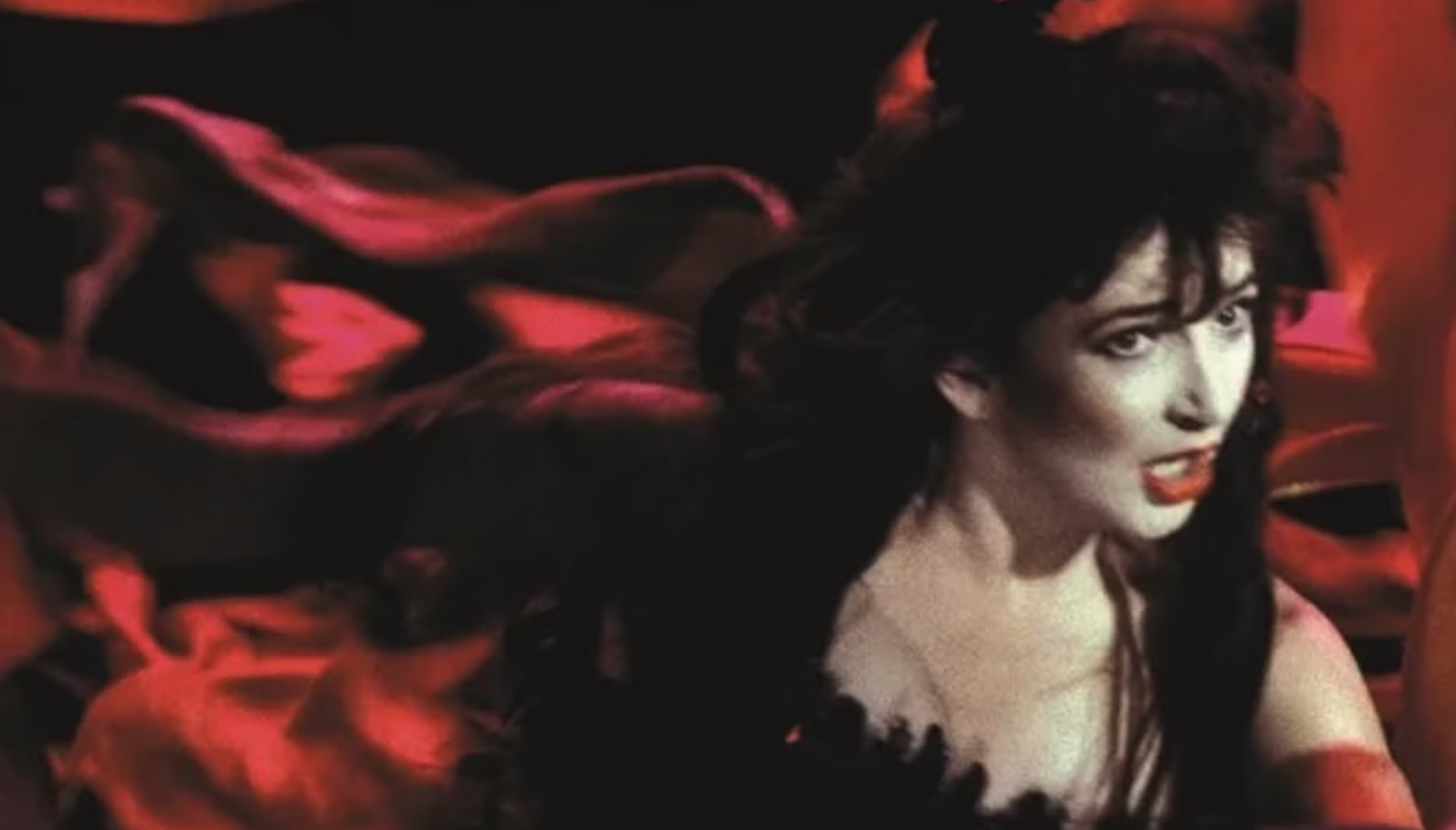A film still of a pale woman with long brown hair (Kate Bush) singing. She's wearing a flowing red garment.