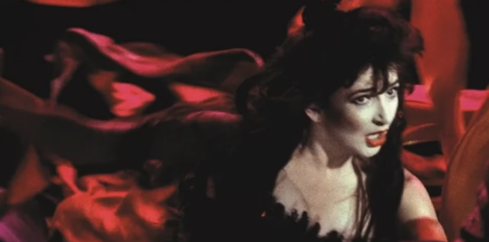 A film still of a pale woman with long brown hair (Kate Bush) singing. She's wearing a flowing red garment.