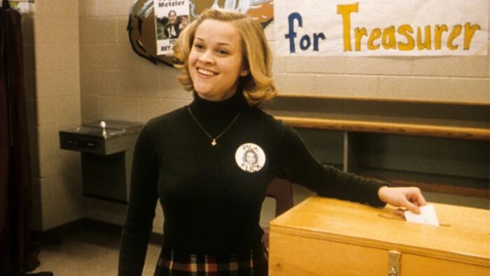 A young woman with a blond bob and a black turtleneck on, standing next to a ballot box.