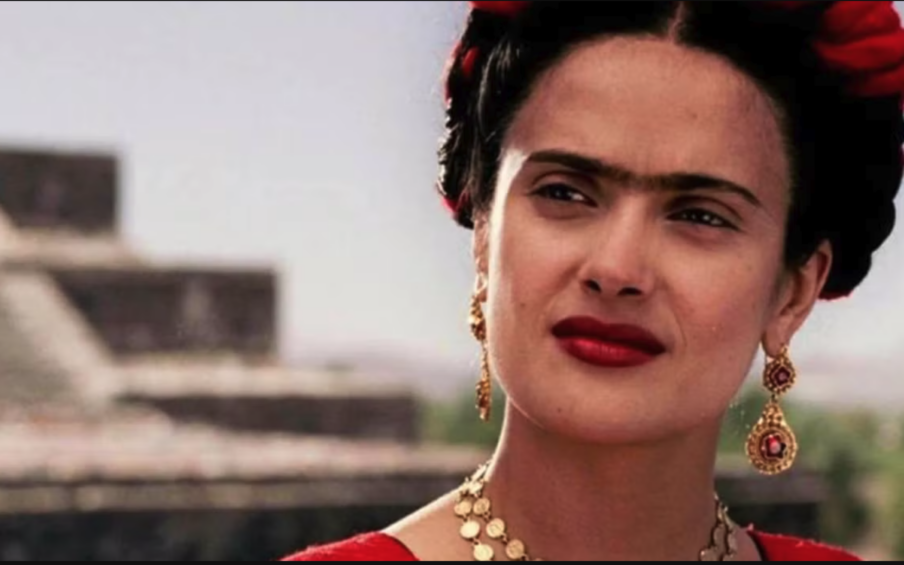 Salma Hayek dressed as artist Frida Kahlo standing in front of a pyramid.