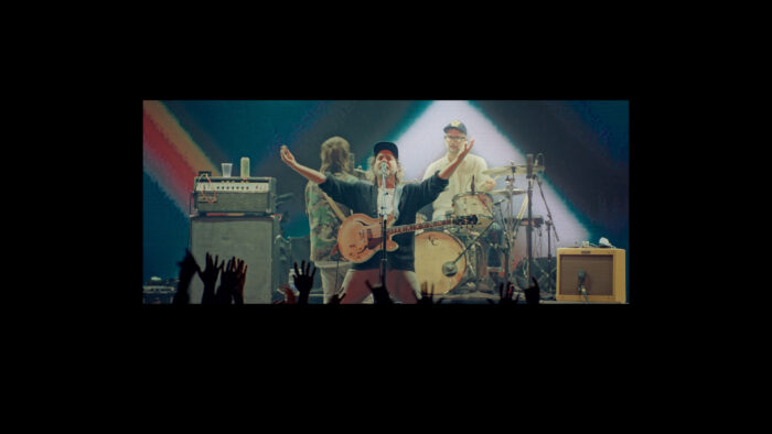 Film still of a rock band on stage. The guitarist at the front center is holding his arms out and has his mouth wide open.