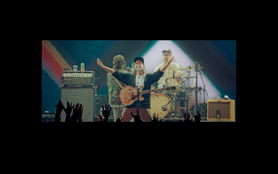Film still of a rock band on stage. The guitarist at the front center is holding his arms out and has his mouth wide open.