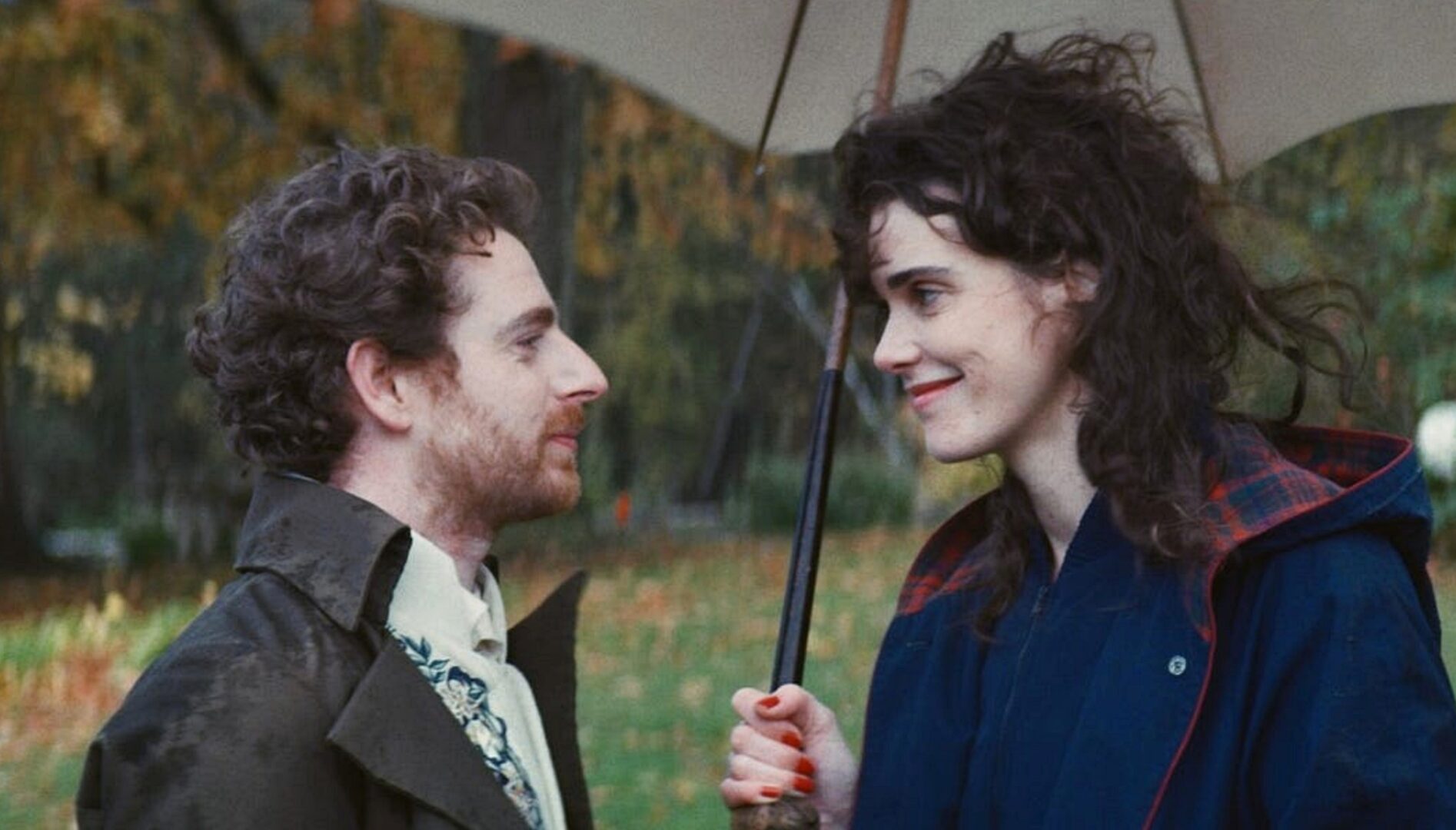 A man and a woman, both with curly brown hair, looking at each other under an umbrella