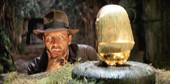 A man in a brown leather jacket and brown brimmed hat looking cautiously at a gold item on a pedestal in a cave.