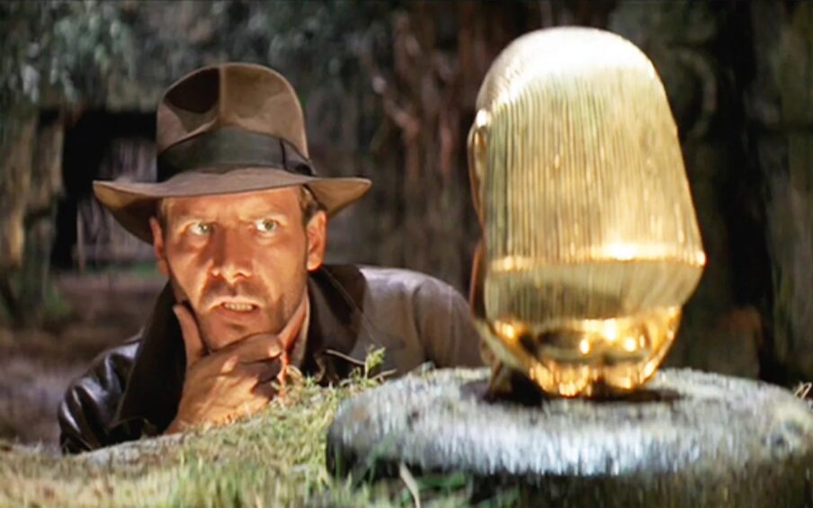 A man in a brown leather jacket and brown brimmed hat looking cautiously at a gold item on a pedestal in a cave.
