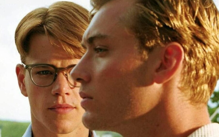 A blond man with eyeglasses (Matt Damon) in the background staring towards a blond man (Jude Law) looking to the side.