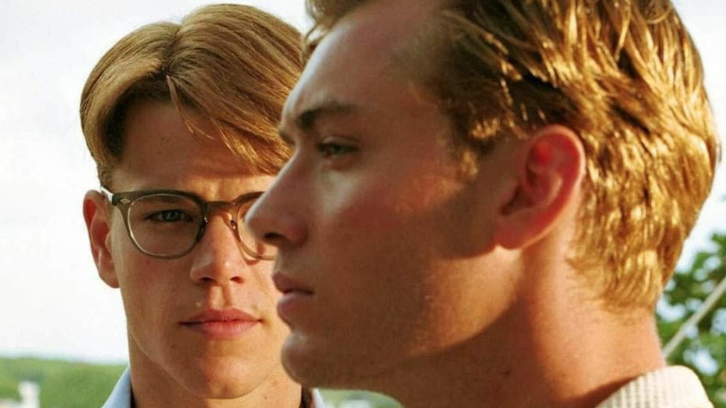A blond man with eyeglasses (Matt Damon) in the background staring towards a blond man (Jude Law) looking to the side.