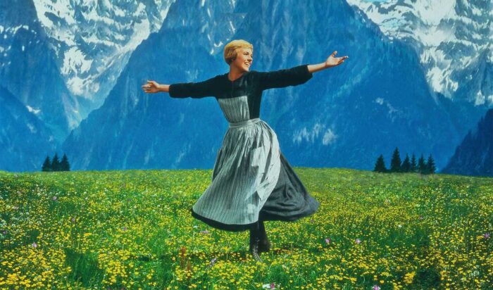 A woman with short blond hair in a dress and apron twirling on green grass with a backdrop of mountains.