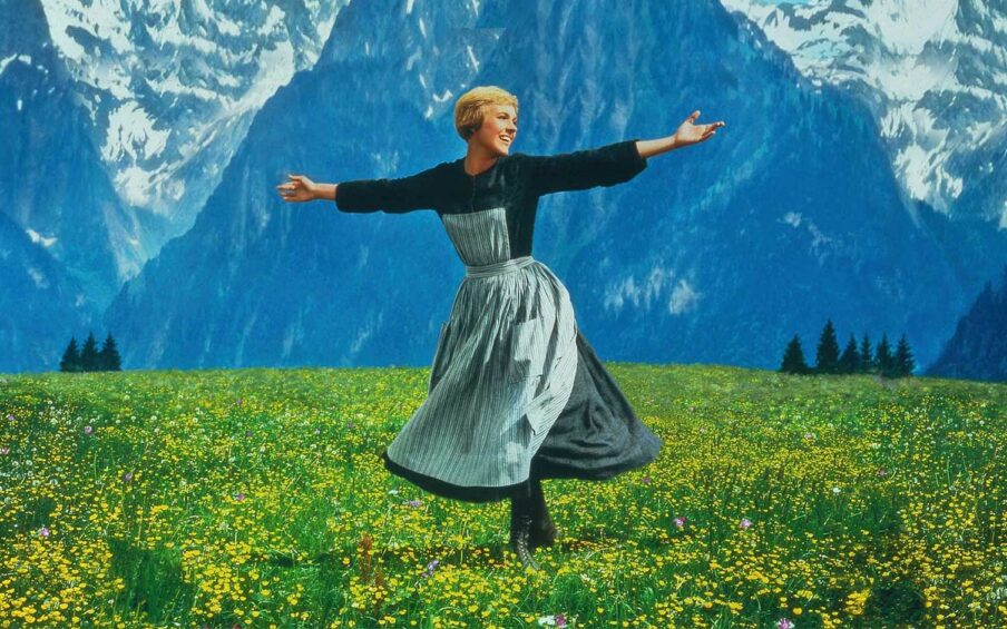 A woman with short blond hair in a dress and apron twirling on green grass with a backdrop of mountains.