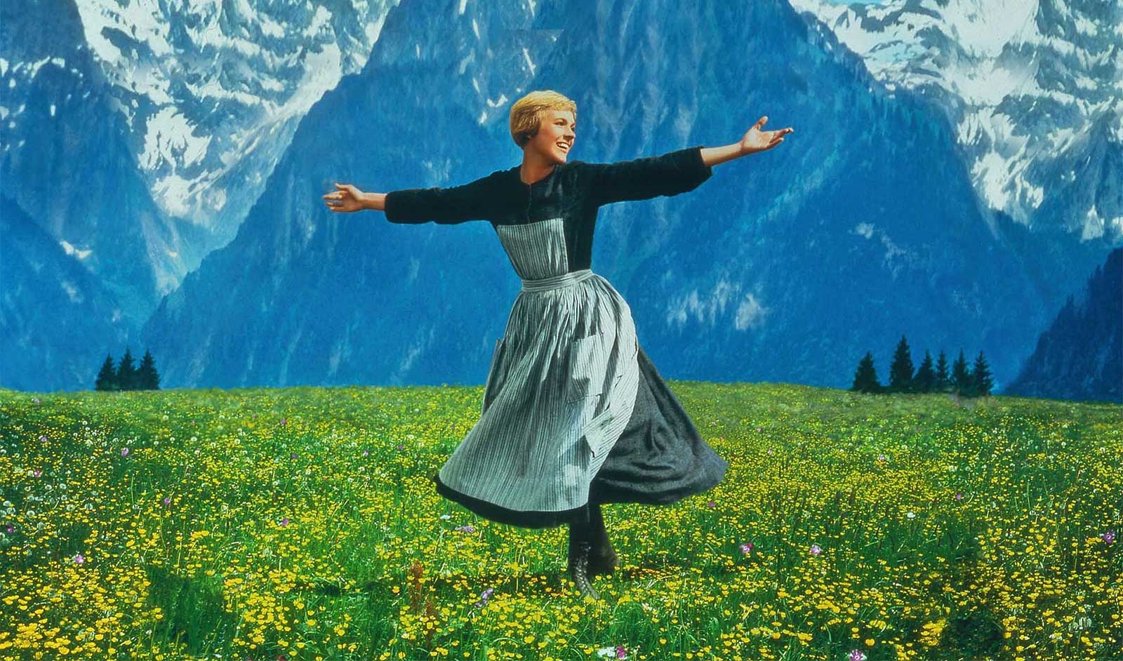 A woman with short blond hair in a dress and apron twirling on green grass with a backdrop of mountains.