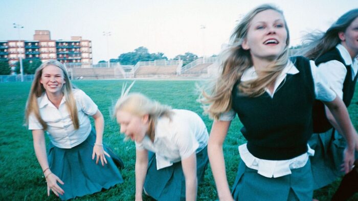 Four blond girls in school uniforms laughing and playing on an expanse of grass.