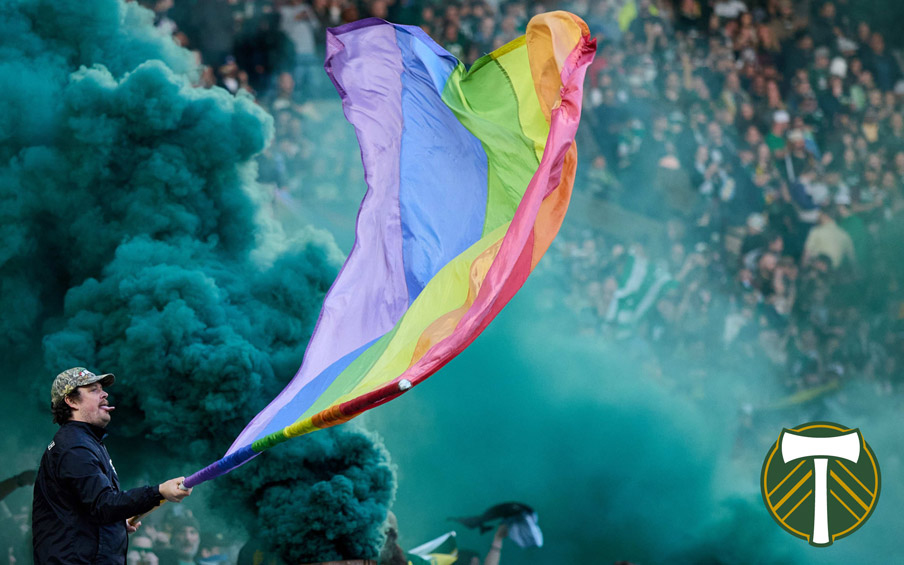 A person waving a large rainbow flag in front of a cloud of blue smoke with the Timbers logo overlayed.