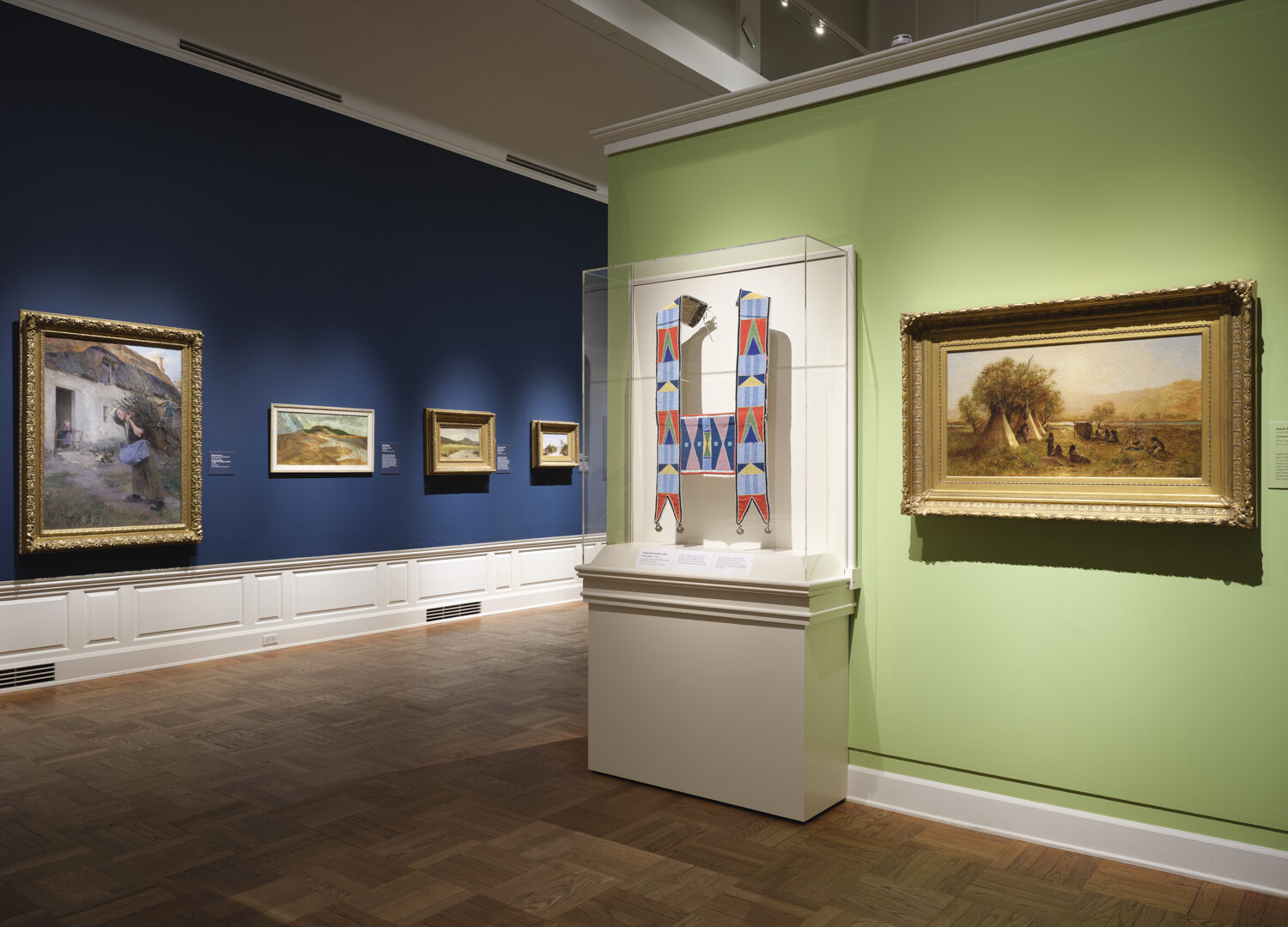 Photo of a gallery with dark blue and light green walls with paintings hung and a case with a Native beadwork object.