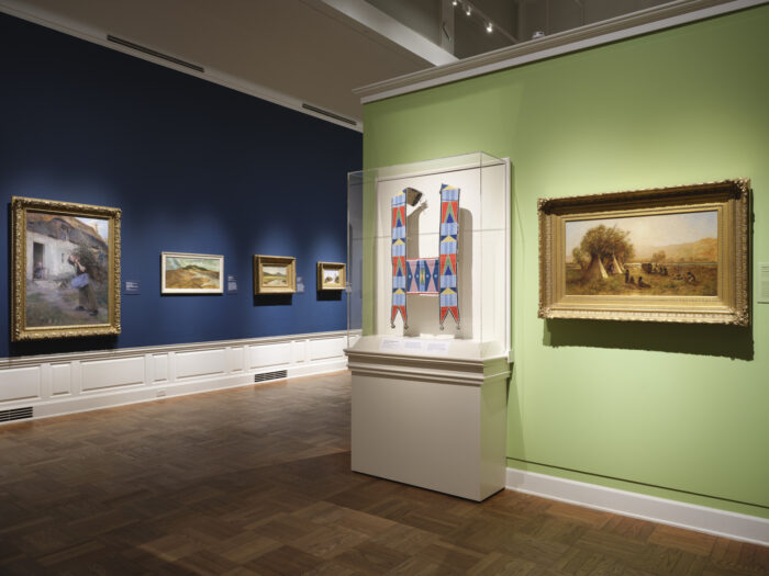 Photo of a gallery with dark blue and light green walls with paintings hung and a case with a Native beadwork object.